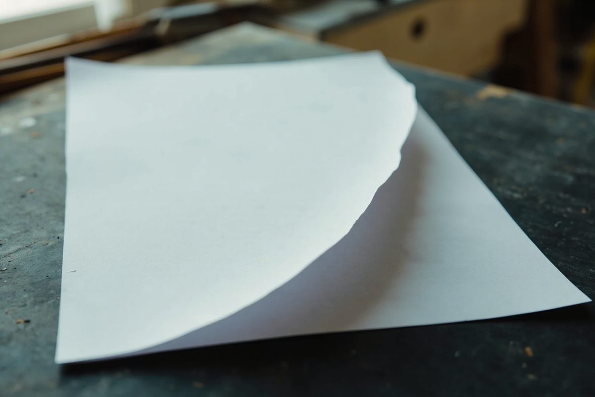 A sheet of thin plain white card on a dark bench, one corner lifted in a soft curved bend with the paper broken along the raised edge.