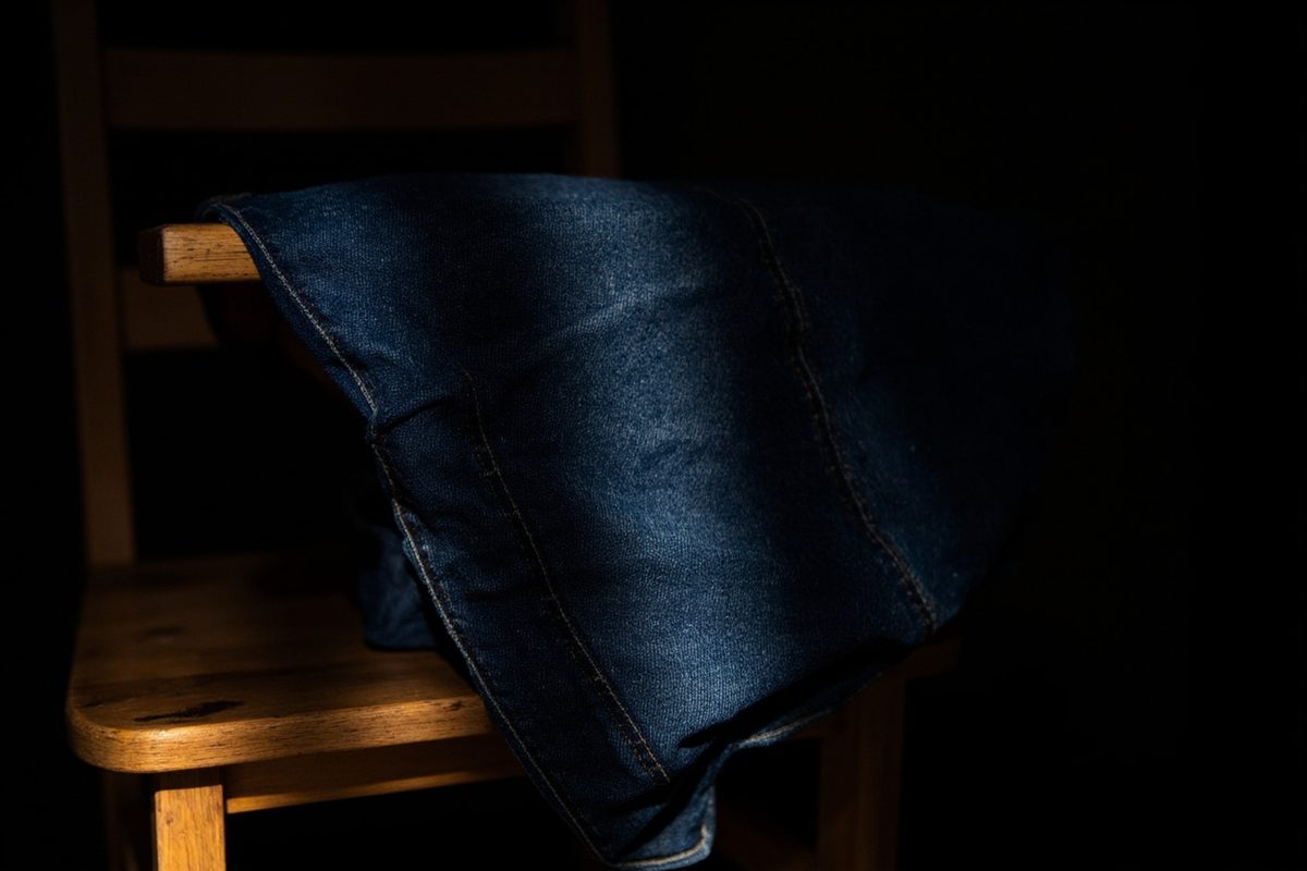 Plain indigo jeans draped over a wooden chair back under warm side light, with worn lighter areas showing across the thigh.
