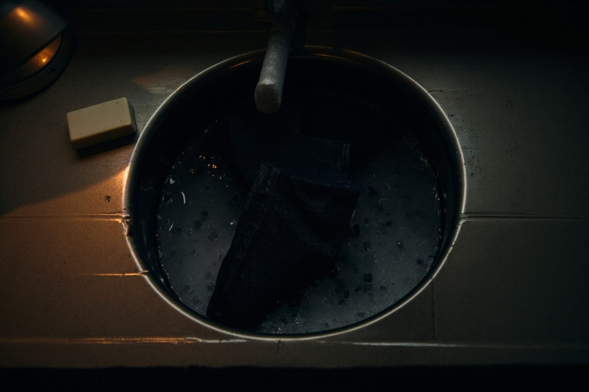 Plain dark jeans submerged in a sink of cool water beside a plain bar of soap, with no suds.