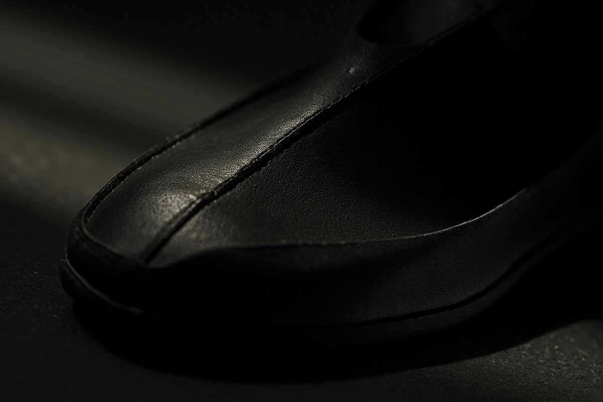A plain dark leather shoe photographed from a low angle under raking light, with deep flex creases across the toe box.