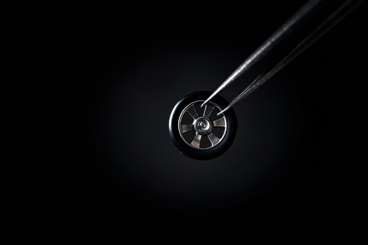 Extreme close-up of a small blank rubber toy car wheel with a plain chrome hub held in tweezers.