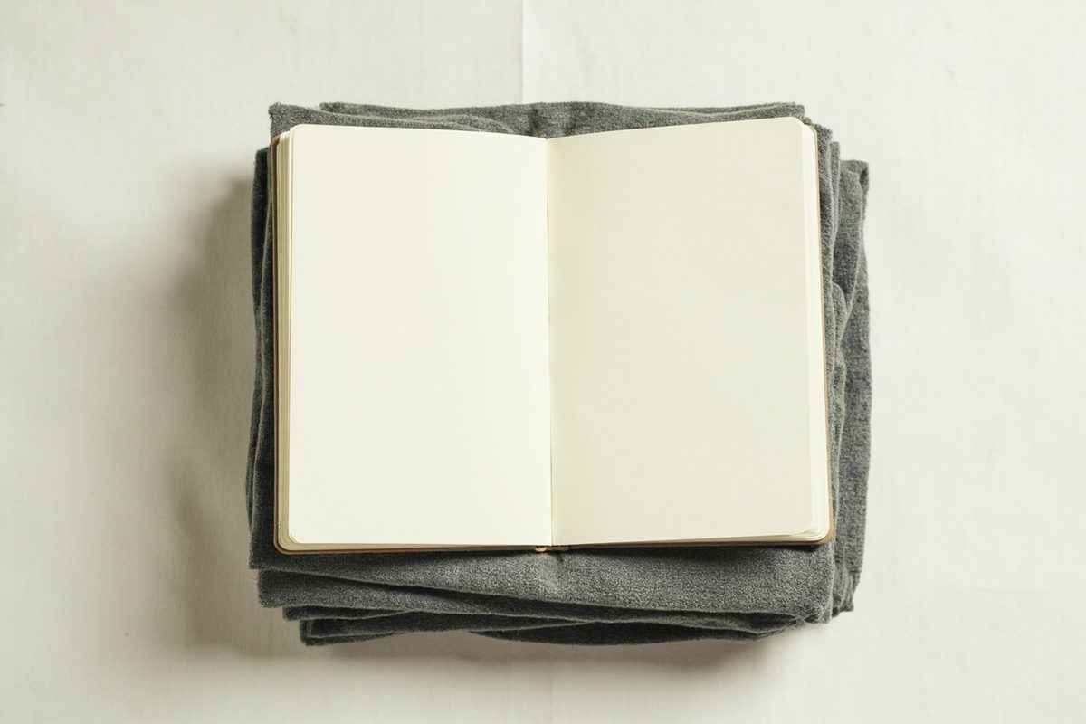 An open notebook with blank pages resting on a neat square stack of plainly folded grey garments.