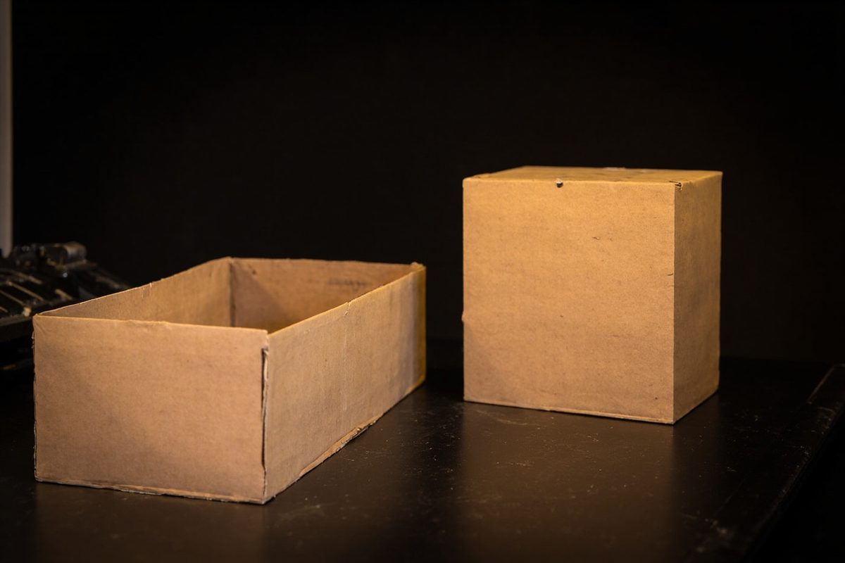 An empty open cardboard tray beside a closed intact cardboard box on a dark bench.