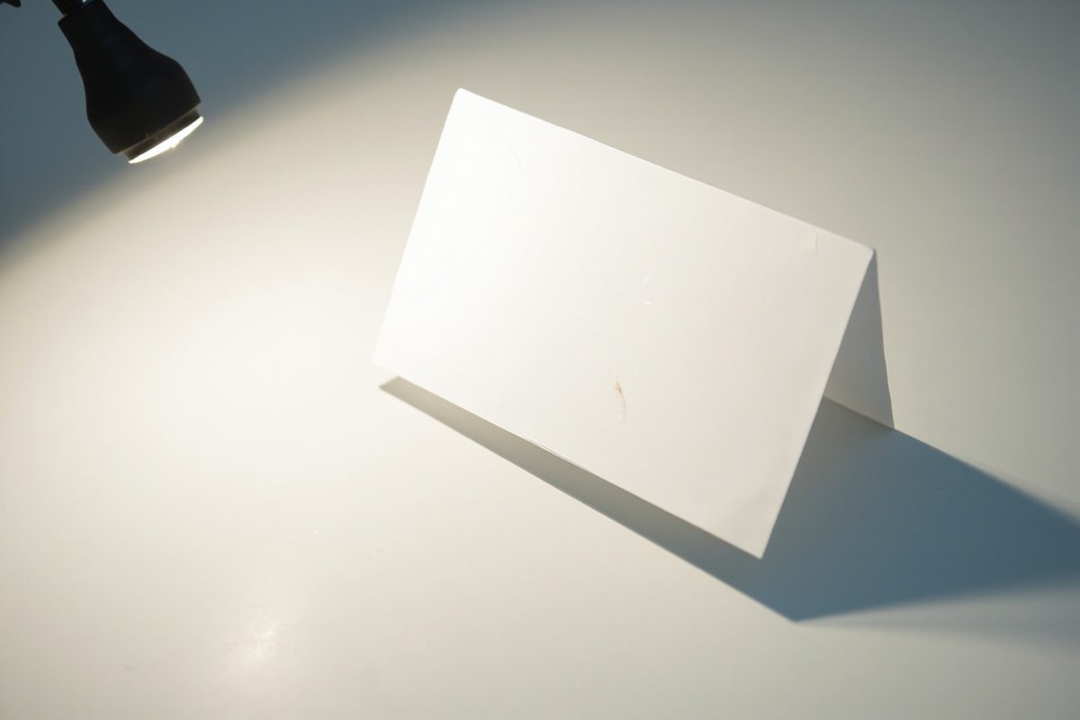 A blank card propped upright under a single desk lamp, the raking light throwing a long shadow and picking up faint surface marks.