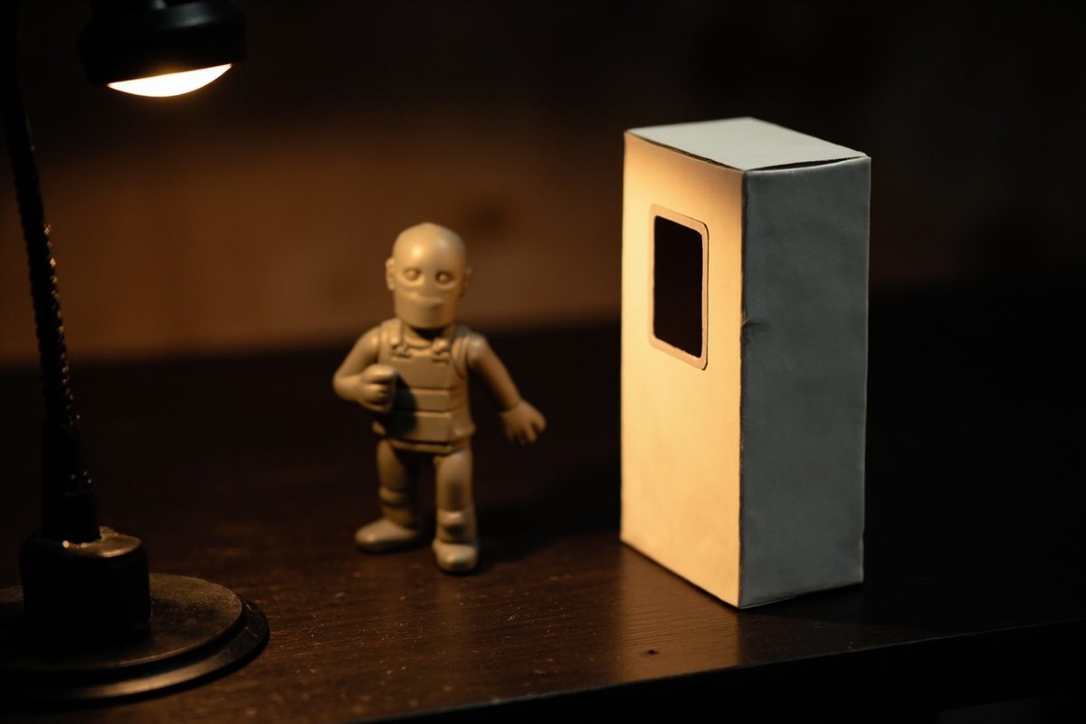 A plain unprinted window box standing on a bench beside a loose figure, lit by a desk lamp.