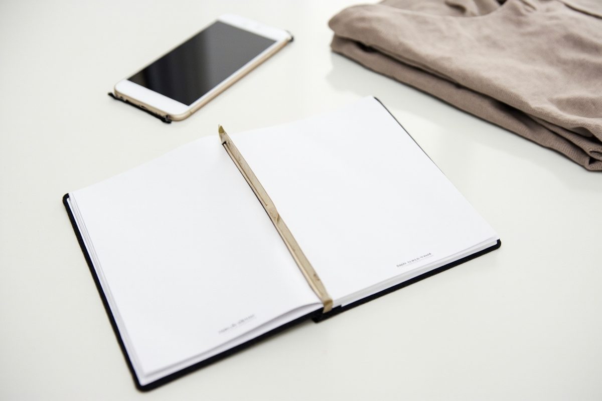 An open blank notebook and pencil on a pale desk, a phone and a folded plain garment beside it.