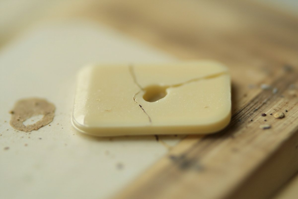Top-down close-up of a pale rigid plastic part with fine hairline cracks running out from the edge of a small pin hole.